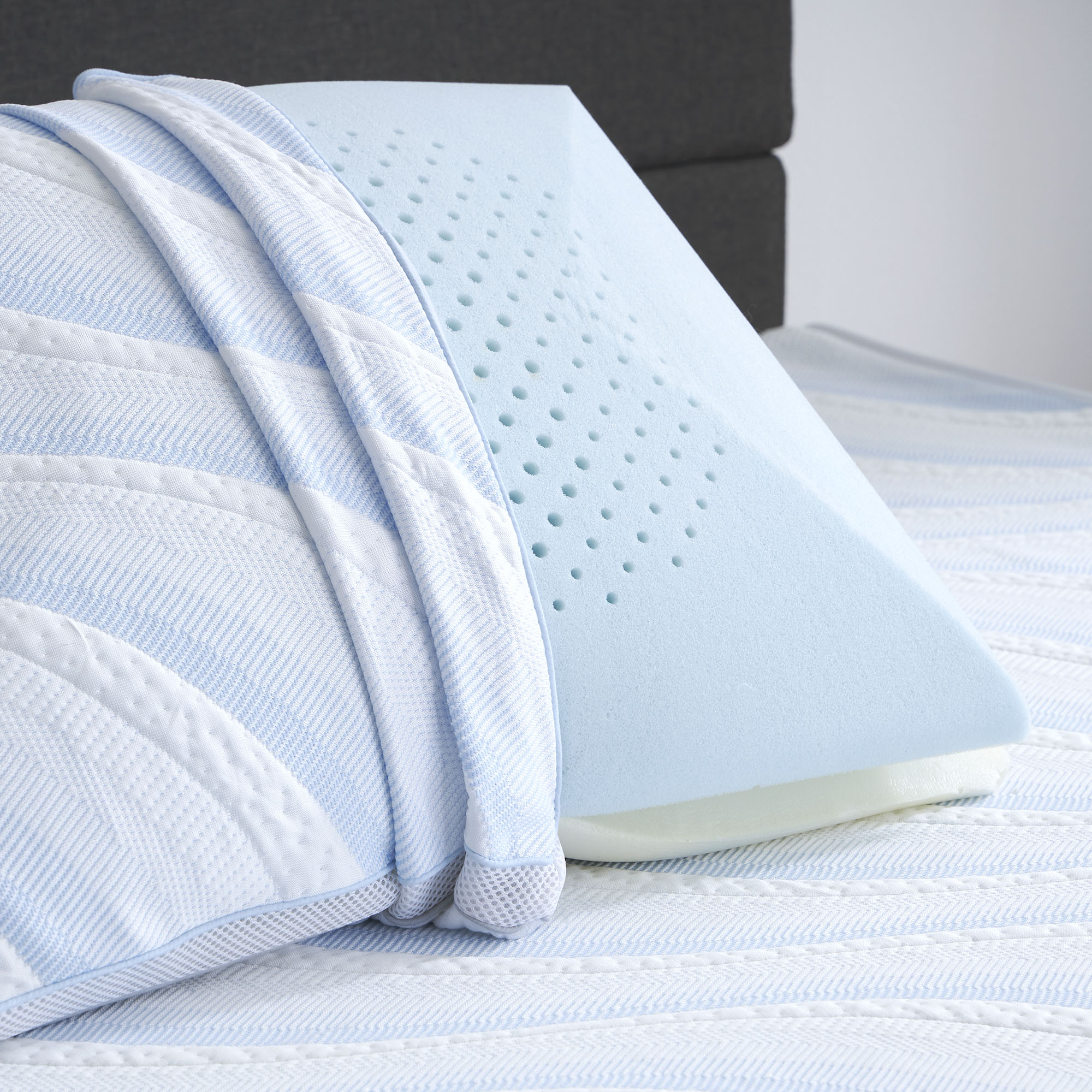 A Comfort Tech Dual Comfort Foam Pillow with a blue cover sits upright on a white and light blue striped bed, featuring small ventilation holes and a textured pattern, with a matching blanket draped over the cooling pillow.