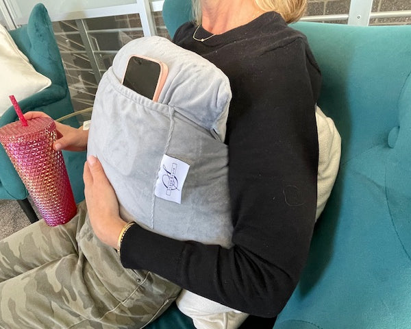 A person relaxes on a teal couch, wearing a black top and camouflage pants, using the Chest Pillow with a pocket to hold their smartphone for comfort and support after breast surgery while holding a pink tumbler.
