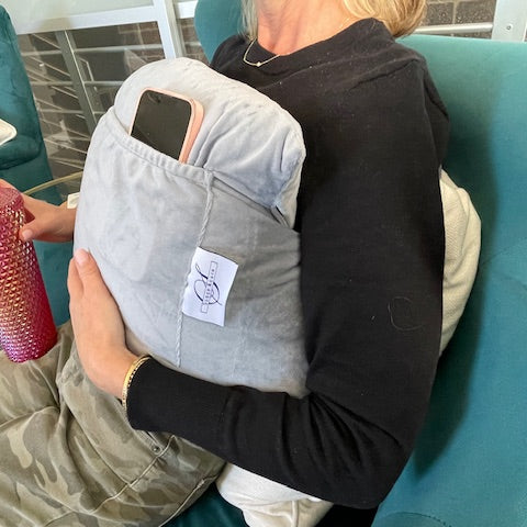 A person relaxes on a teal couch, wearing a black top and camouflage pants, using the Chest Pillow with a pocket to hold their smartphone for comfort and support after breast surgery while holding a pink tumbler.