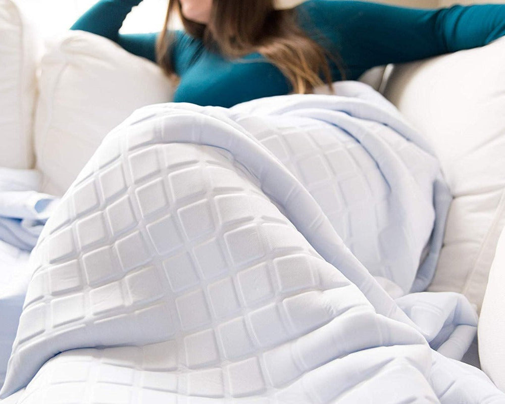 A woman in a teal long-sleeve shirt relaxes on a white couch, covered with the Final Sale: Opulence Glacier Blanket, a cooling and weighted white blanket with a geometric square pattern. Her face is partially out of frame.
