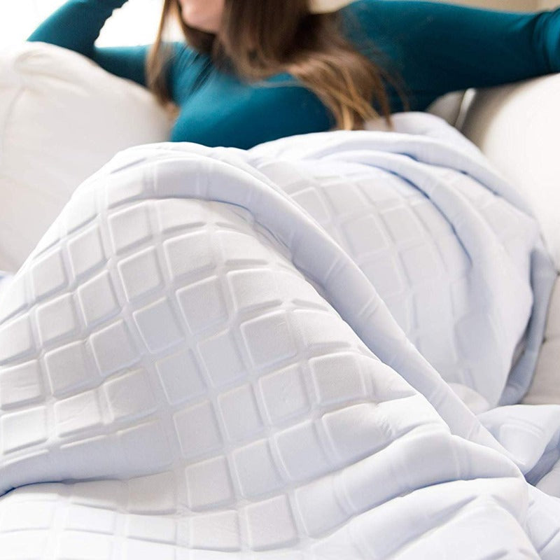 A woman in a teal long-sleeve shirt relaxes on a white couch, covered with the Final Sale: Opulence Glacier Blanket, a cooling and weighted white blanket with a geometric square pattern. Her face is partially out of frame.