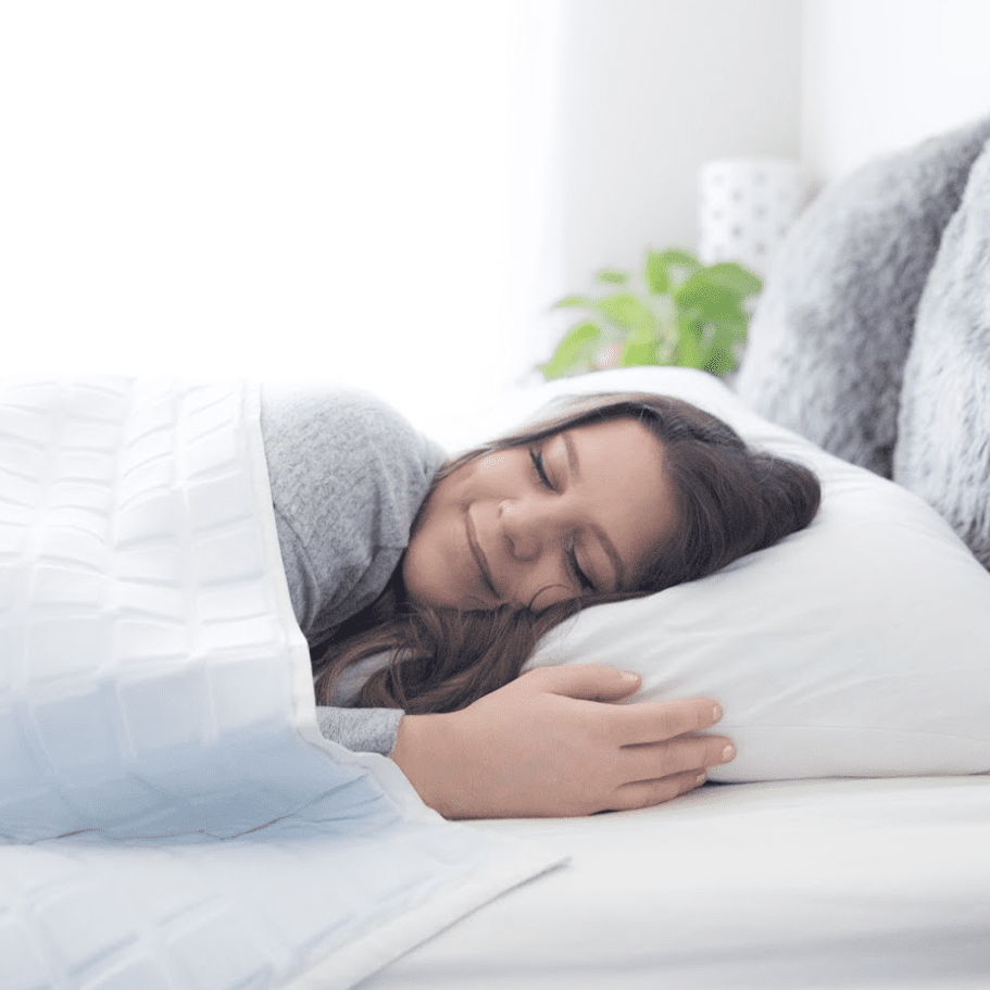 A young woman with long brown hair peacefully sleeps on a white bed, hugging a pillow and wrapped in the Final Sale: Opulence Glacier Blanket | Cooling & Weighted. Soft light fills the cozy, neatly decorated bedroom.