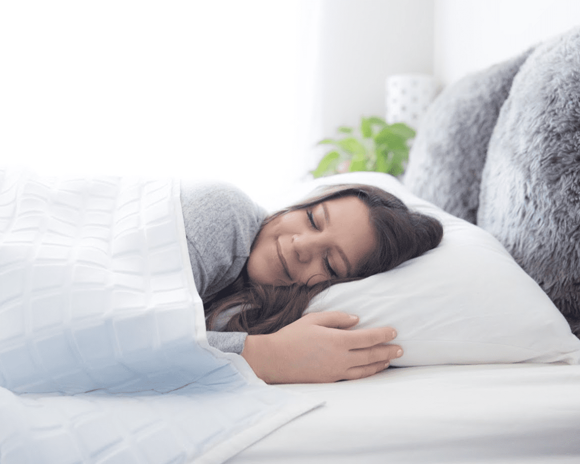 A young woman with long brown hair peacefully sleeps on a white bed, hugging a pillow and wrapped in the Final Sale: Opulence Glacier Blanket | Cooling & Weighted. Soft light fills the cozy, neatly decorated bedroom.