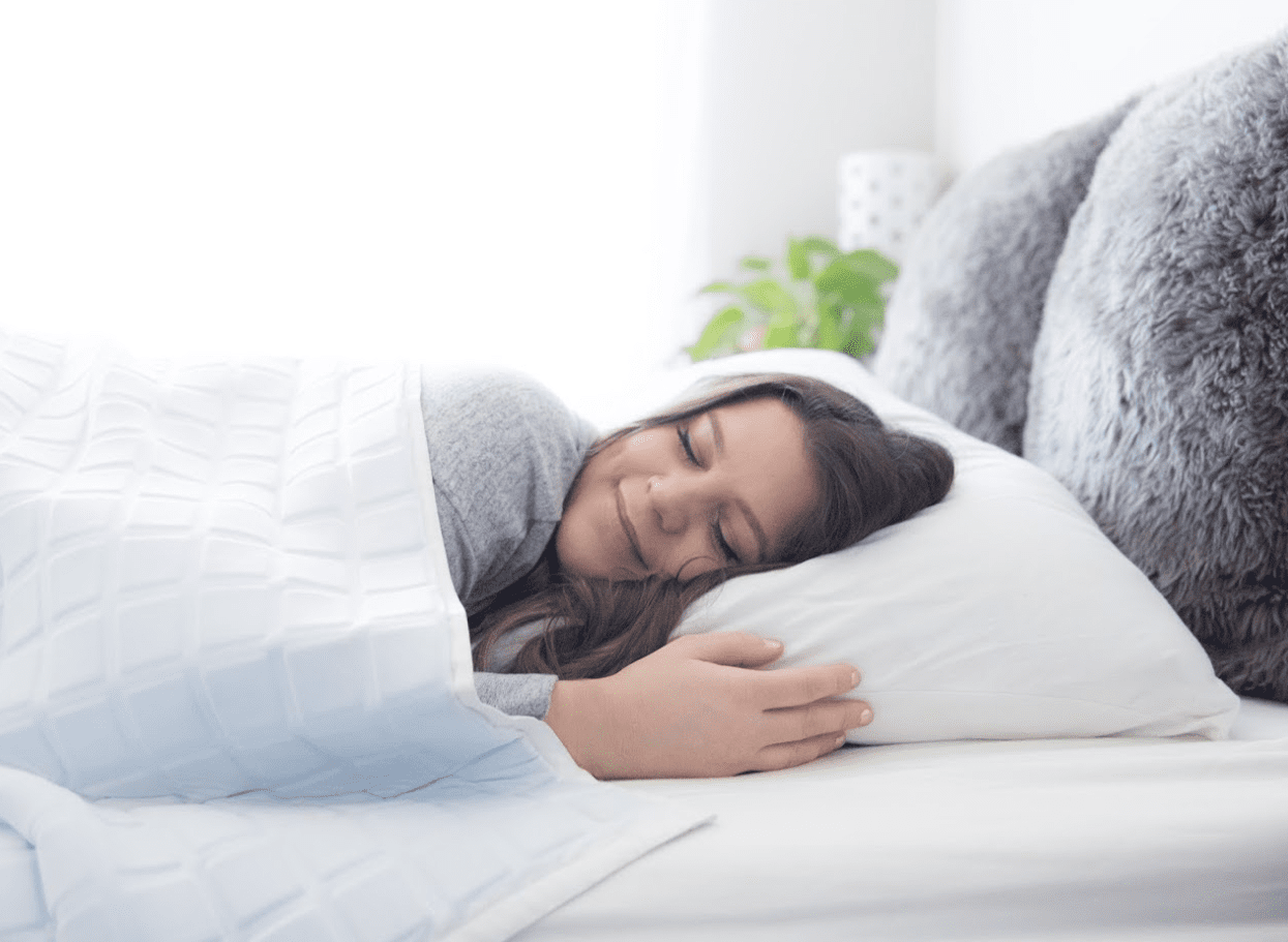 A young woman with long brown hair peacefully sleeps on a white bed, hugging a pillow and wrapped in the Final Sale: Opulence Glacier Blanket | Cooling & Weighted. Soft light fills the cozy, neatly decorated bedroom.