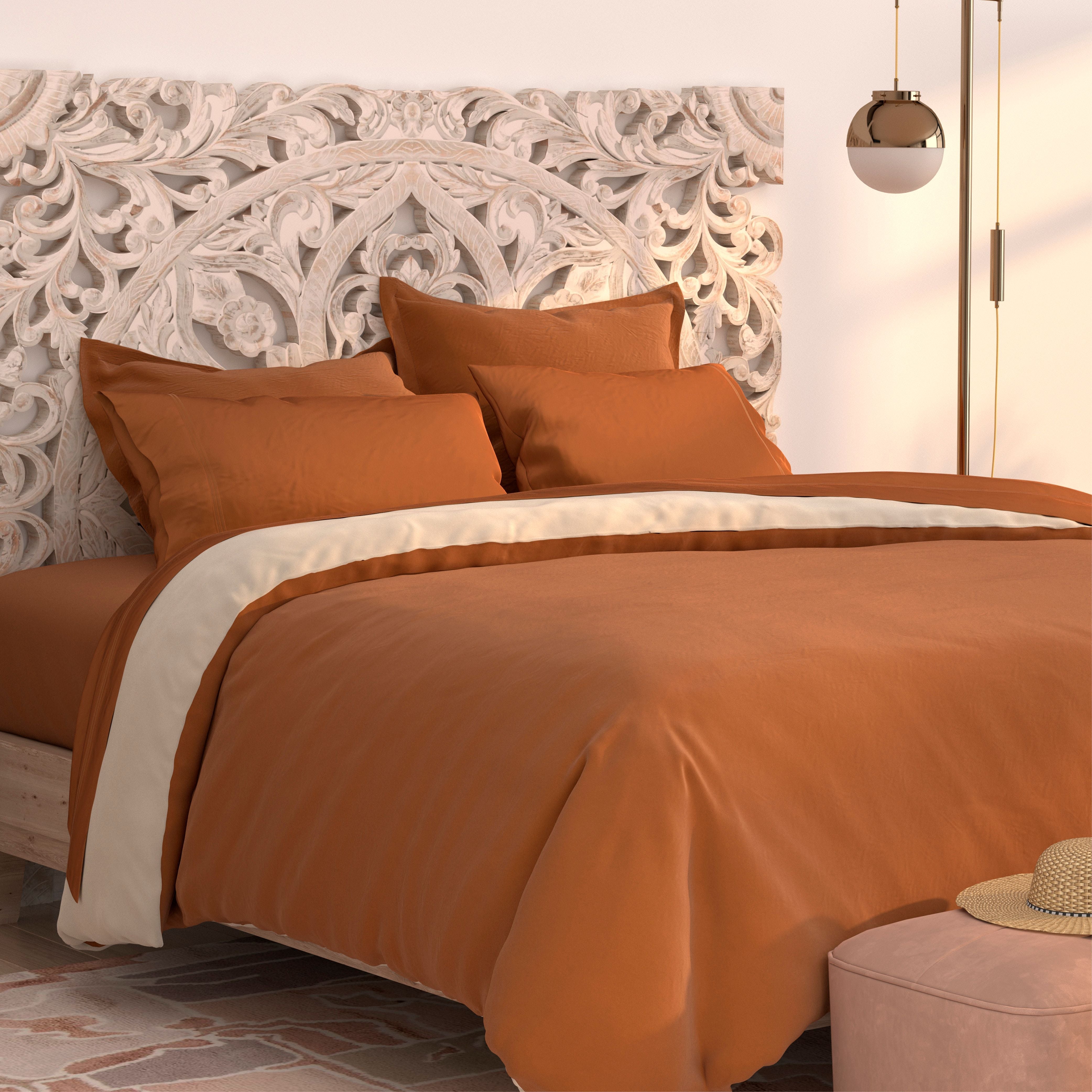 A bed features the PureCare Duvet Cover | Soft Touch Bamboo in burnt orange and cream against an ornate carved wooden headboard. A round pendant light and plush ottoman complete the softly lit, modern bedroom.
