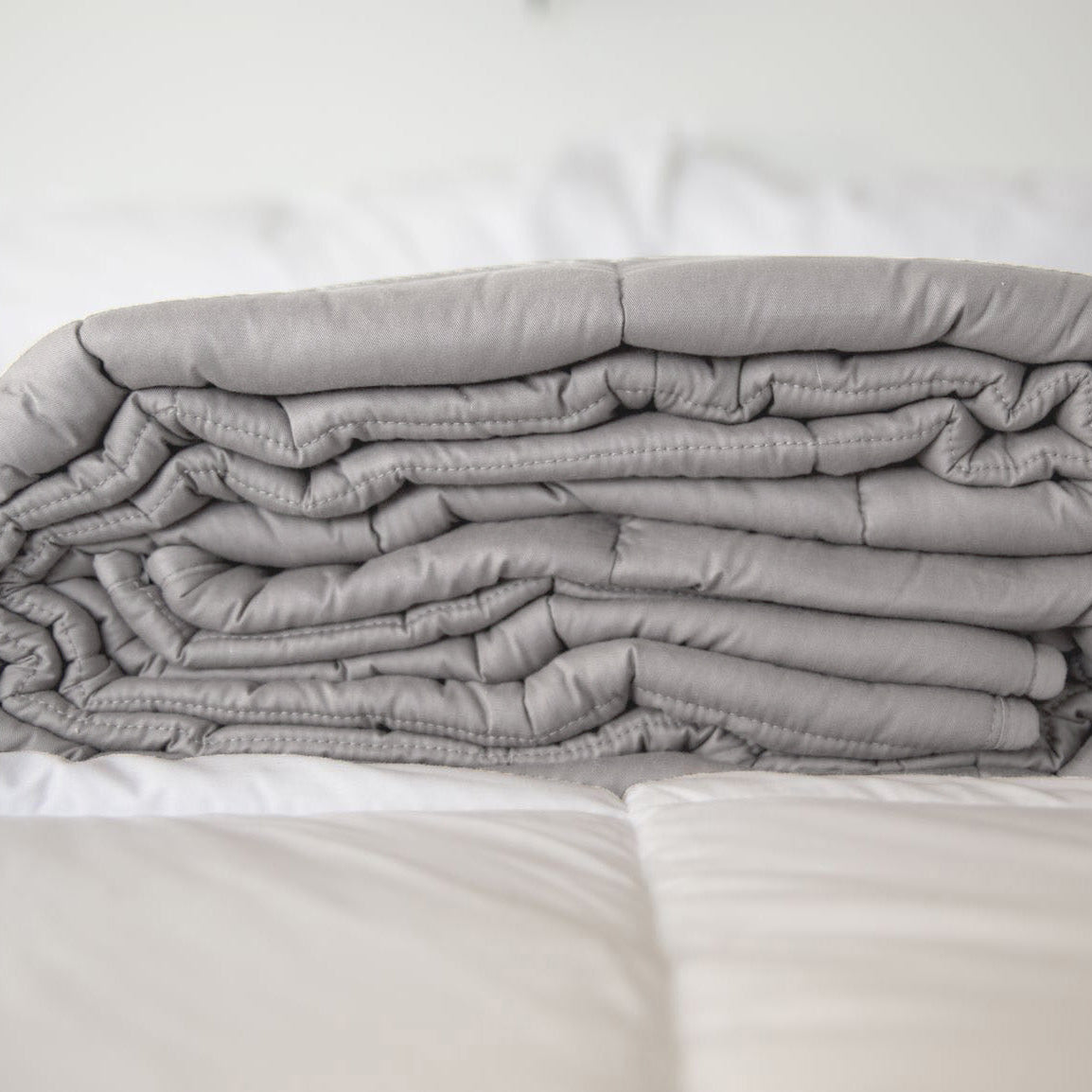 A neatly folded Pillowtex Weighted Blanket in gray rests atop a white, quilted bedspread in a softly lit room.