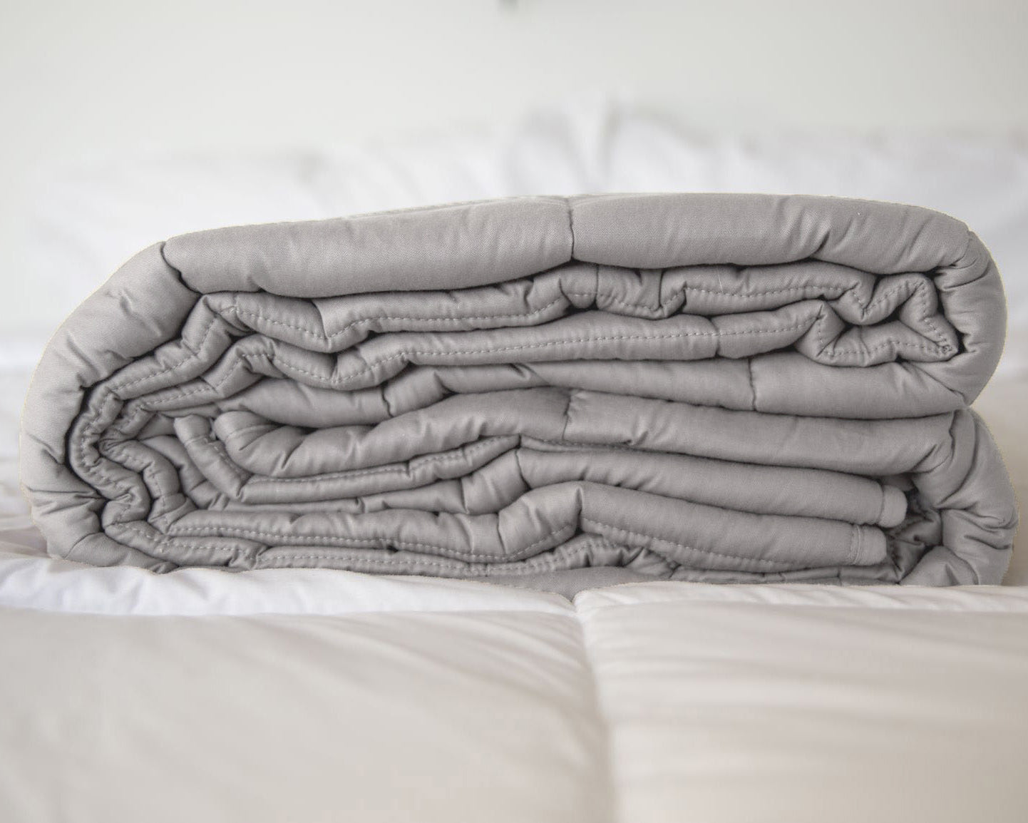 A neatly folded Pillowtex Weighted Blanket in gray rests atop a white, quilted bedspread in a softly lit room.
