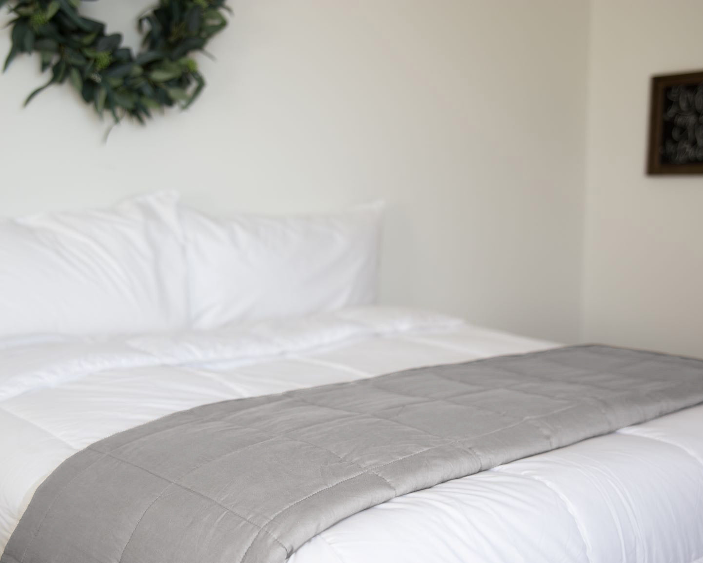 A neatly made bed with a Pillowtex Weighted Blanket folded at the foot and white bedding. A leafy wreath decorates the wall above, while a framed chalkboard hangs on the adjacent wall.