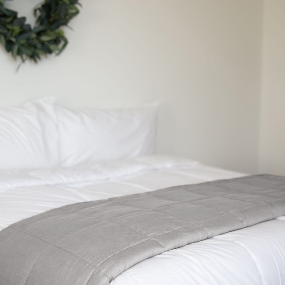 A neatly made bed with a Pillowtex Weighted Blanket folded at the foot and white bedding. A leafy wreath decorates the wall above, while a framed chalkboard hangs on the adjacent wall.