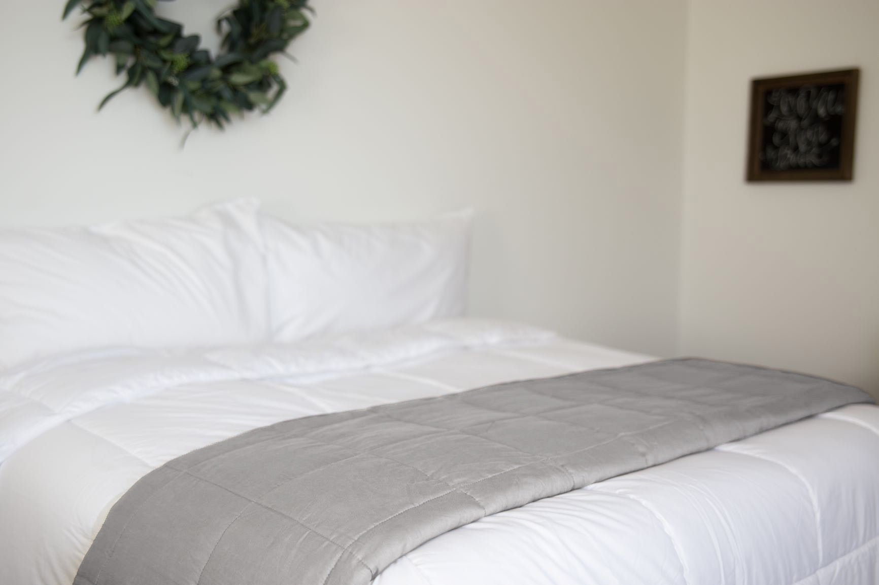 A neatly made bed with a Pillowtex Weighted Blanket folded at the foot and white bedding. A leafy wreath decorates the wall above, while a framed chalkboard hangs on the adjacent wall.