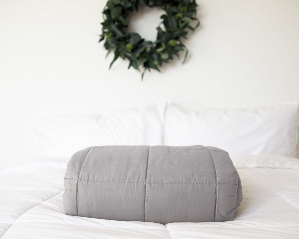 A neatly folded Pillowtex Weighted Blanket in gray rests on a white bed with white pillows, while a green leafy wreath decorates the white wall in the background.