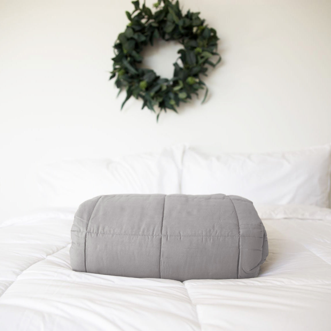 A neatly folded Pillowtex Weighted Blanket in gray rests on a white bed with white pillows, while a green leafy wreath decorates the white wall in the background.