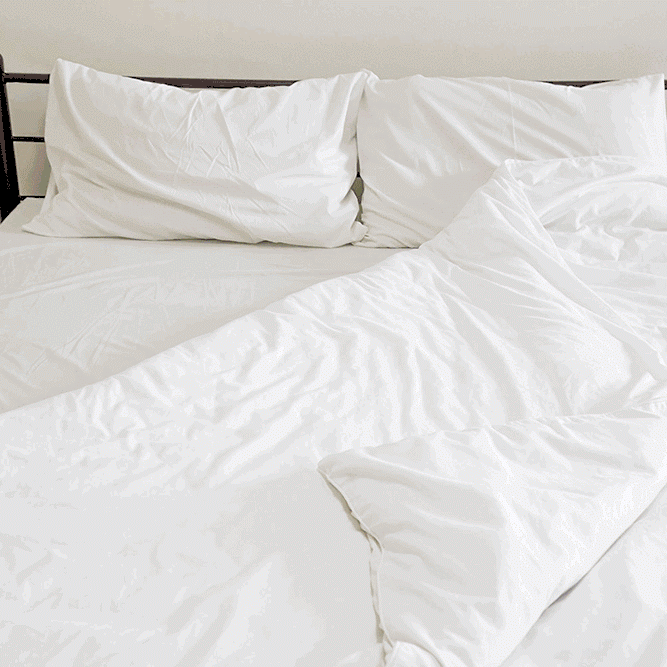A neatly made bed features The Twovet Comforter | Couples Comforter "The Duvet for Two," a white sheet, and two white pillows—one slightly tilted. The blanket is partly folded back to show smooth bedding on a simple dark frame against a plain wall.