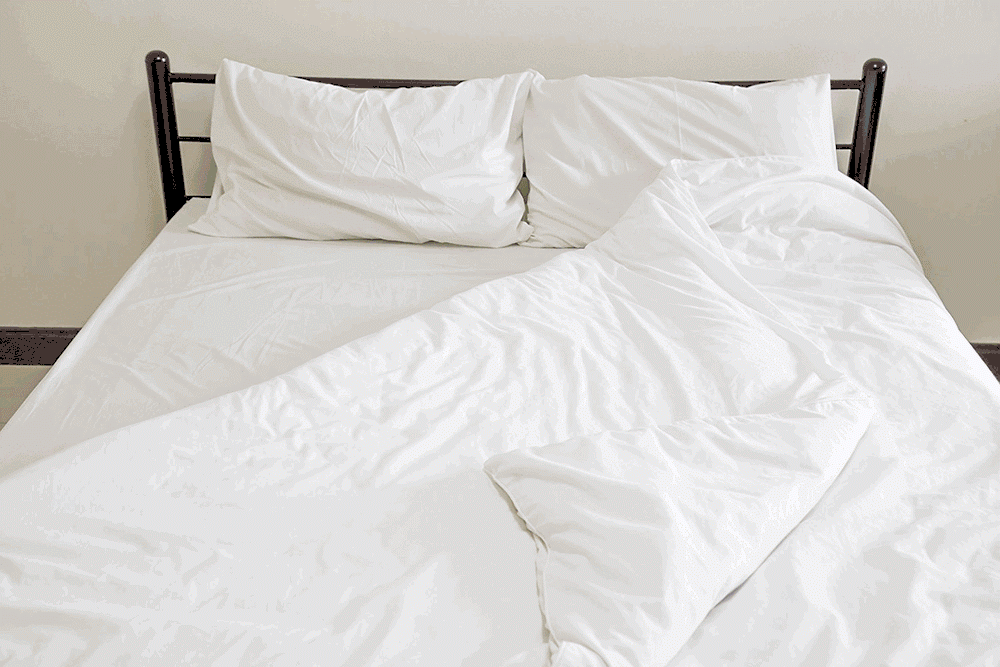 A neatly made bed features The Twovet Comforter | Couples Comforter "The Duvet for Two," a white sheet, and two white pillows—one slightly tilted. The blanket is partly folded back to show smooth bedding on a simple dark frame against a plain wall.