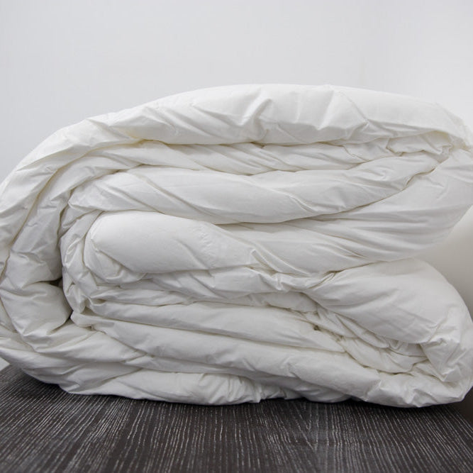 The Twovet Comforter | Couples Comforter "The Duvet for Two," a white, hypoallergenic comforter, is fluffy, neatly rolled, and placed on a dark wooden surface with a plain white wall in the background.