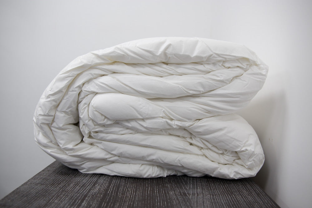 The Twovet Comforter | Couples Comforter "The Duvet for Two," a white, hypoallergenic comforter, is fluffy, neatly rolled, and placed on a dark wooden surface with a plain white wall in the background.