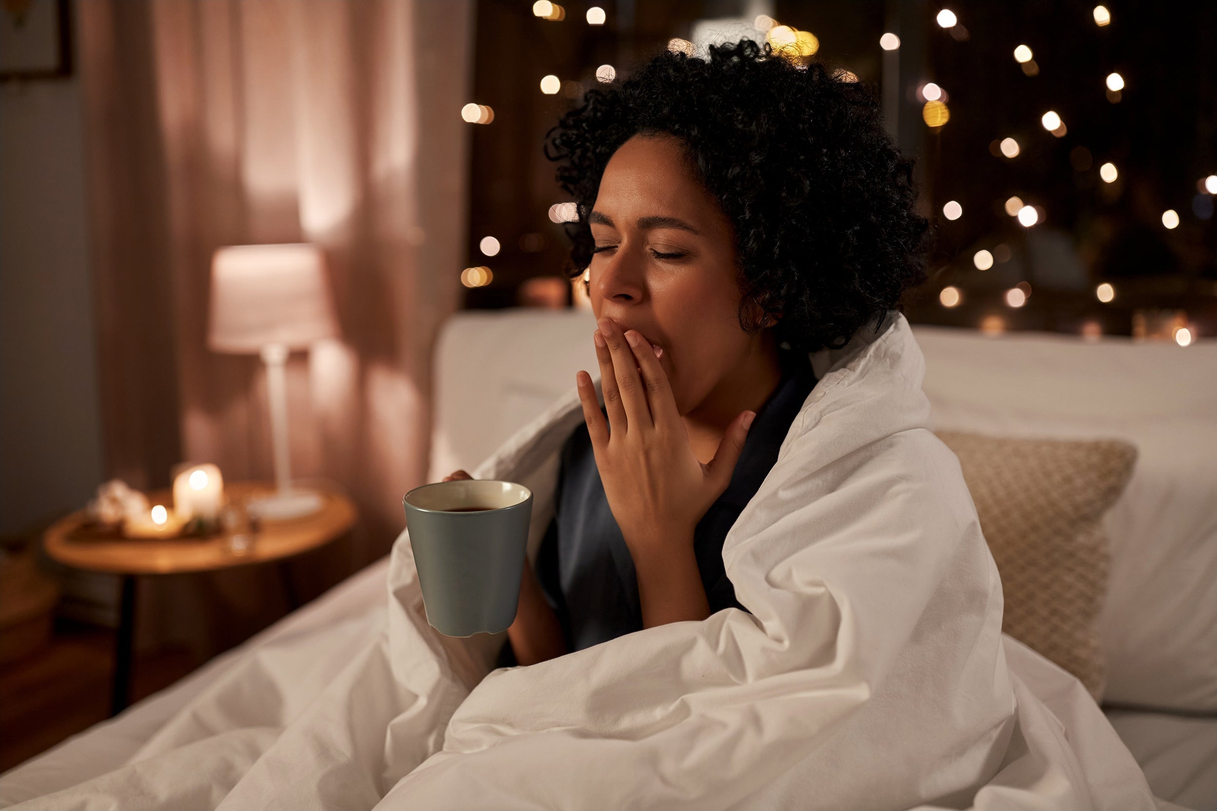 A woman yawns in bed with a cup of coffee. Learn the 10-3-2-1-0 sleep rule: stop caffeine 10 hours before bed, food 3 hours, work 2 hours, screens 1 hour, and hit snooze 0 times for better sleep.