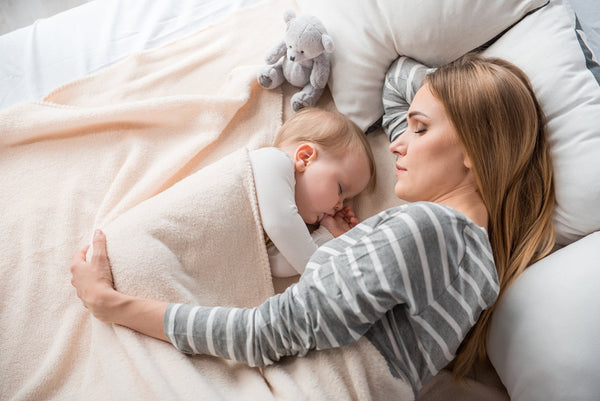 A mother and her newborn baby rest in bed. Sleeping after a C-section can be challenging. Learn more about real solutions for how to sleep after C-section in our new mom survival guide.