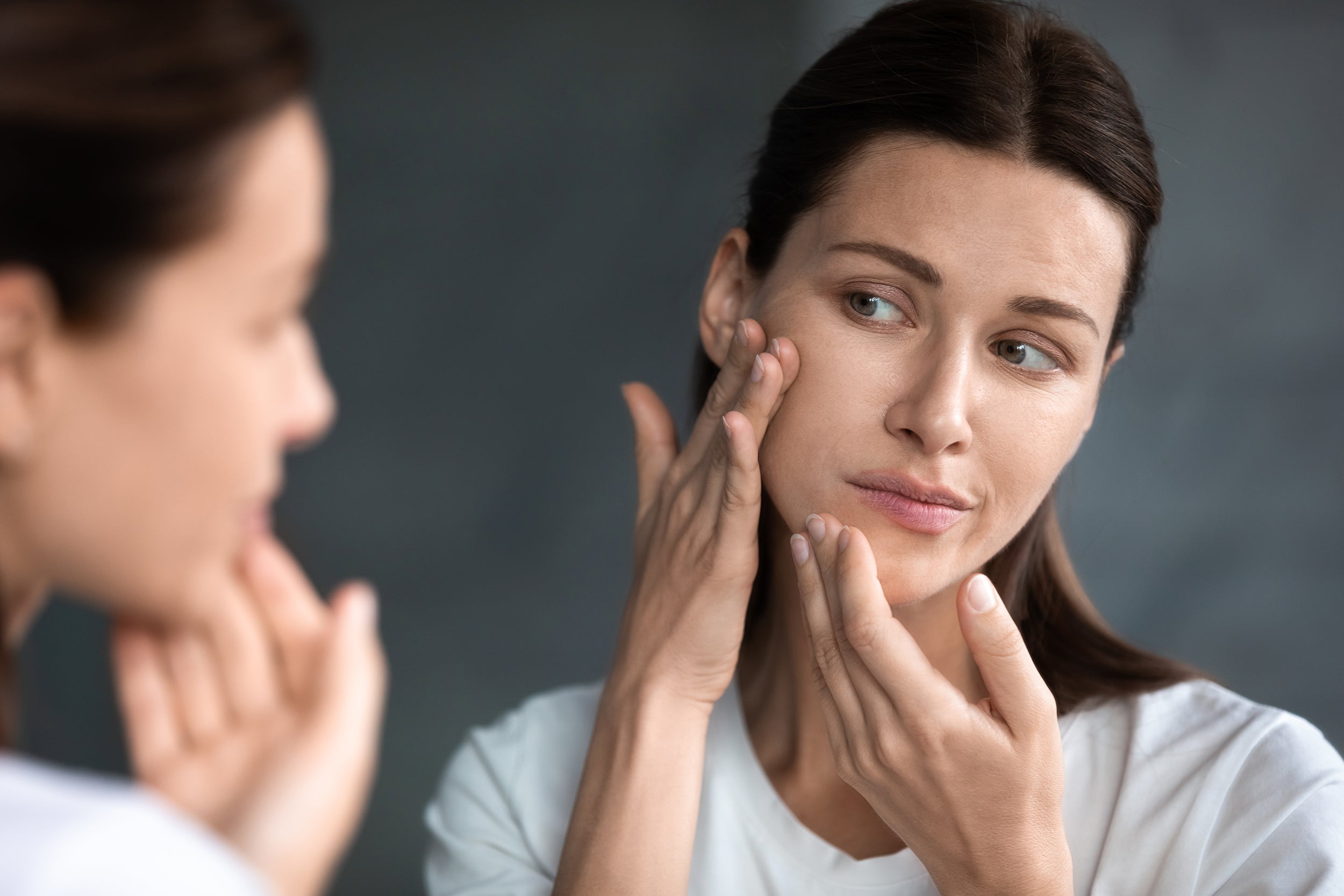 A woman looks at her face in the mirror. Elevated back sleeping at 30–45 degrees is the key to managing swelling after genioplasty. Here's how to maintain it all night without stacked pillows.
