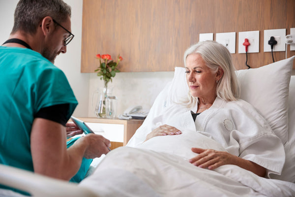 A woman talks to a doctor in a hospital bed. If you're preparing for a tummy tuck, how to sleep after the surgery can make a huge difference in overall healing. Learn more in our complete guide.