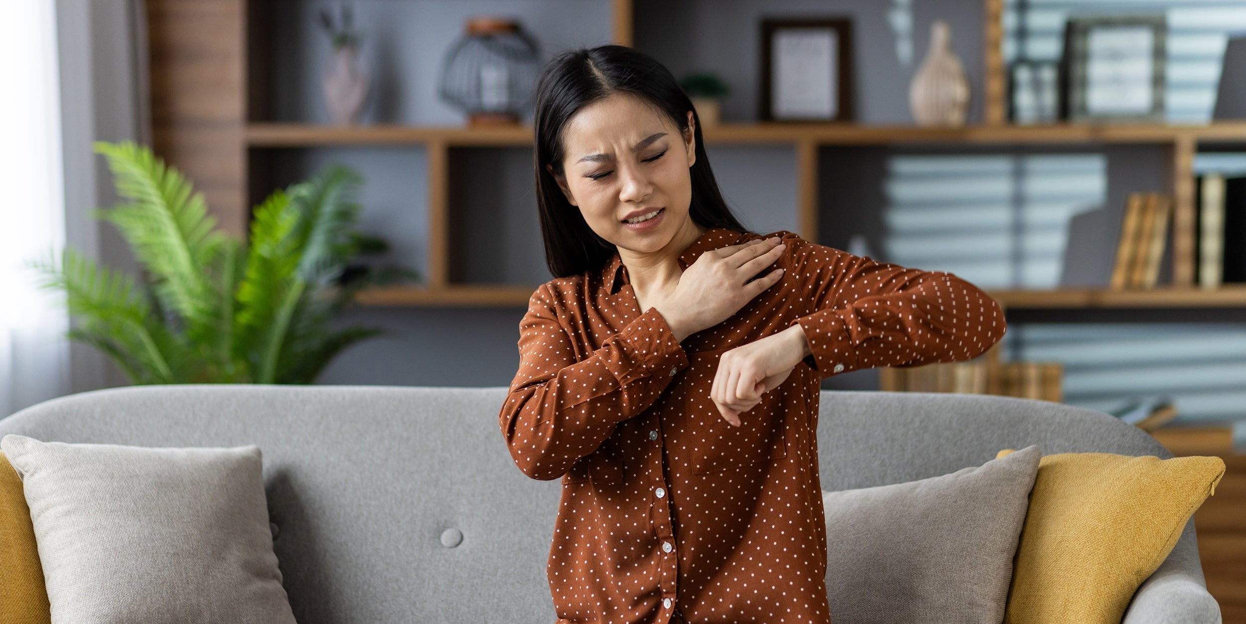 A woman rubs her shoulder in pain. Our ultimate guide to pillows for shoulder pain shares the reasons behind shoulder pain and how to prevent it once and for all with the right shoulder pain pillow.