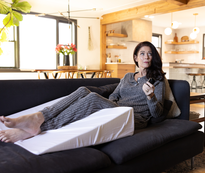 A woman rests on the couch using a single orthopedic wedge pillow for legs from the Sleep Again Pillow System.