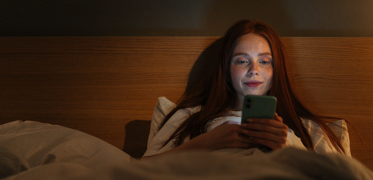 A woman reading her phone in bed. Learn everything you need to know about breast fat transfer recovery, including sleep - one of the most critical recovery pieces.