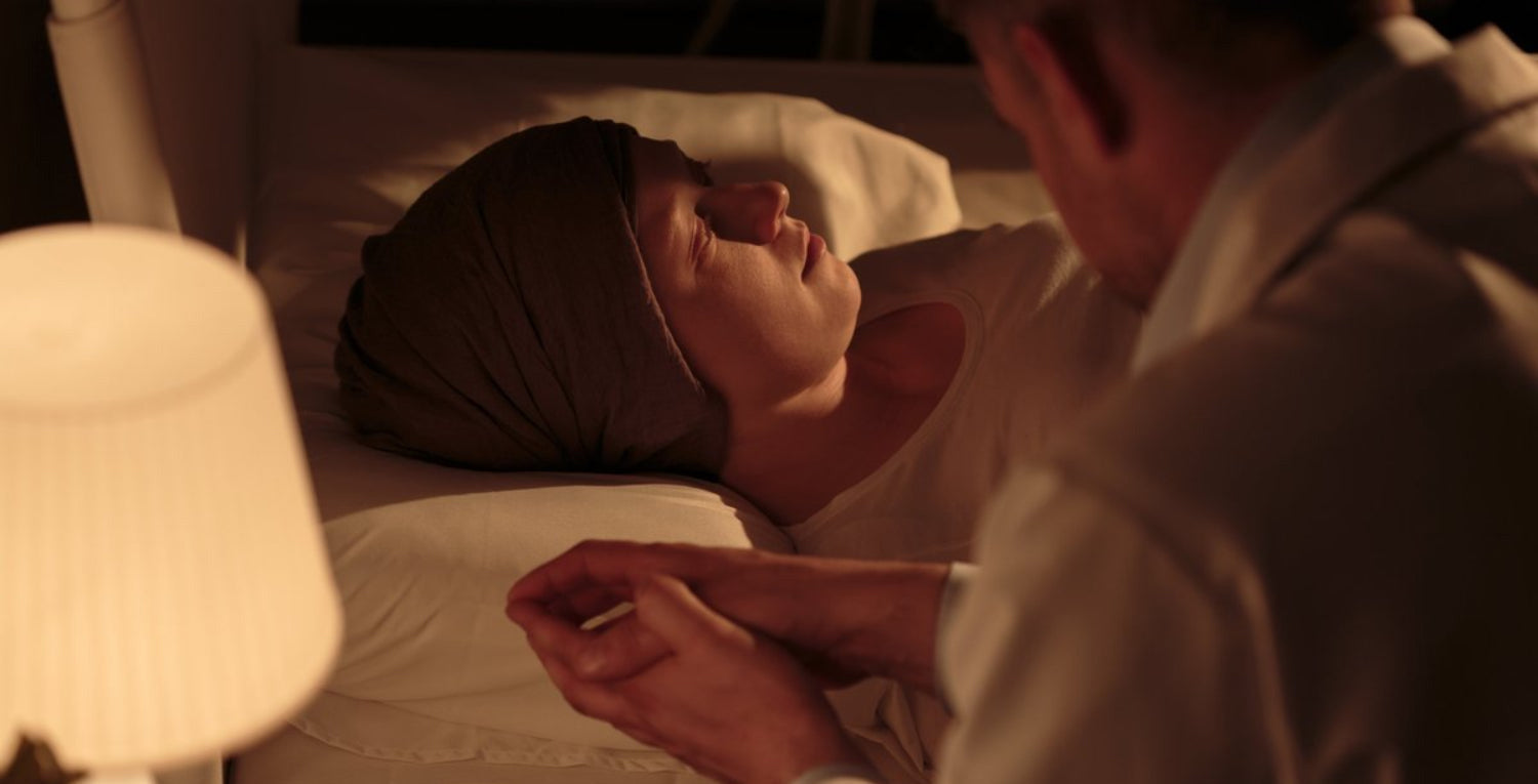 Person resting on their back in bed while a caregiver holds their hand, with a lamp and glass of water on the nightstand.