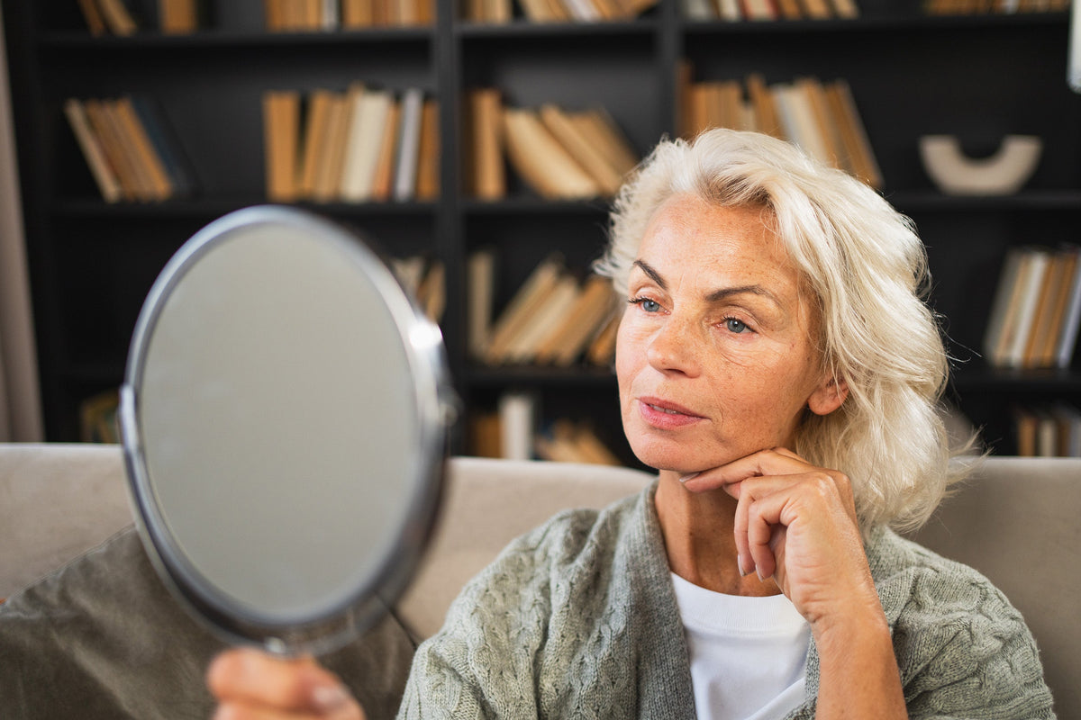 A woman looking in the mirror. Planning a deep plane facelift? Learn how to sleep safely after surgery, how long to stay elevated, and how to set up your recovery space in advance.