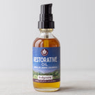 A clear glass bottle with a black pump, labeled Restorative Oil - Arnica, St. John's Wort & Calendula, sits on a light surface. The herb-infused body oil is amber in color and has a purple-blue label with white text.