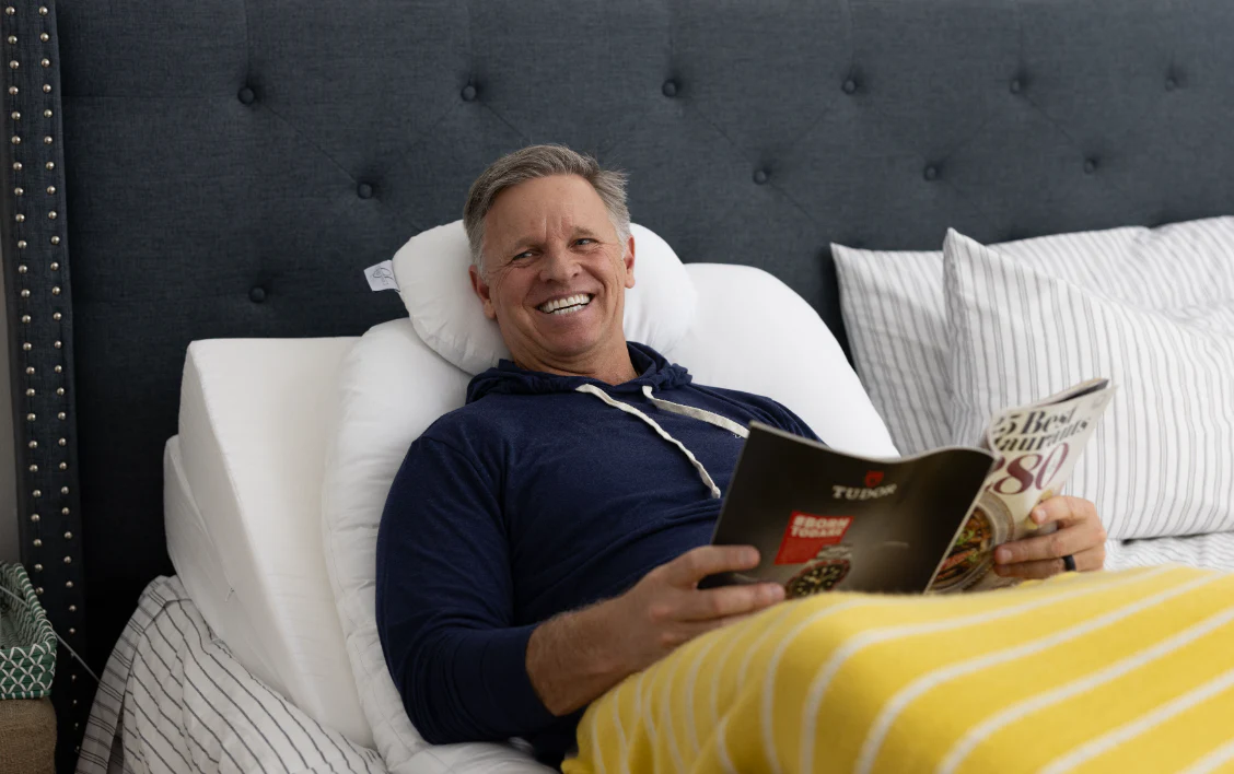 A smiling man in a navy hoodie sits upright in bed against white pillows, covered by a yellow striped blanket, and holds an open magazine. The bed has a dark tufted headboard.