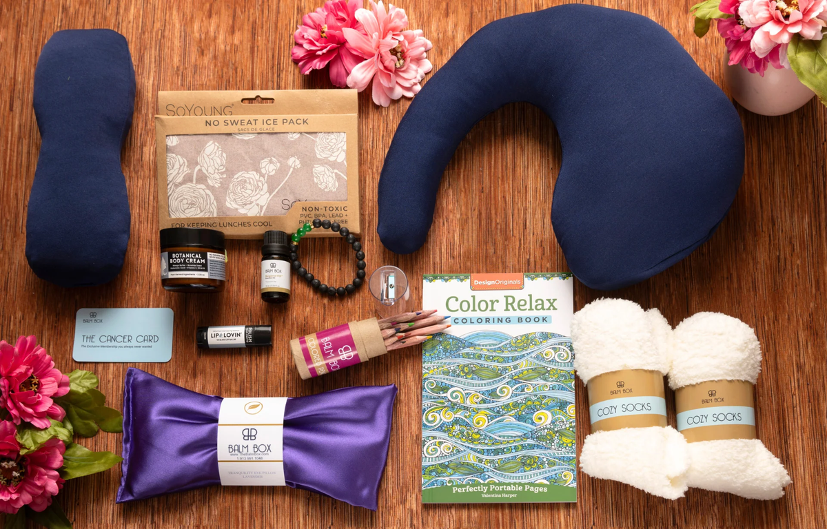 A flat lay of self-care items on a wooden surface, including a neck pillow, eye mask, cozy socks, coloring book with colored pencils, skincare products, a beaded bracelet, and pink flowers for decoration.
