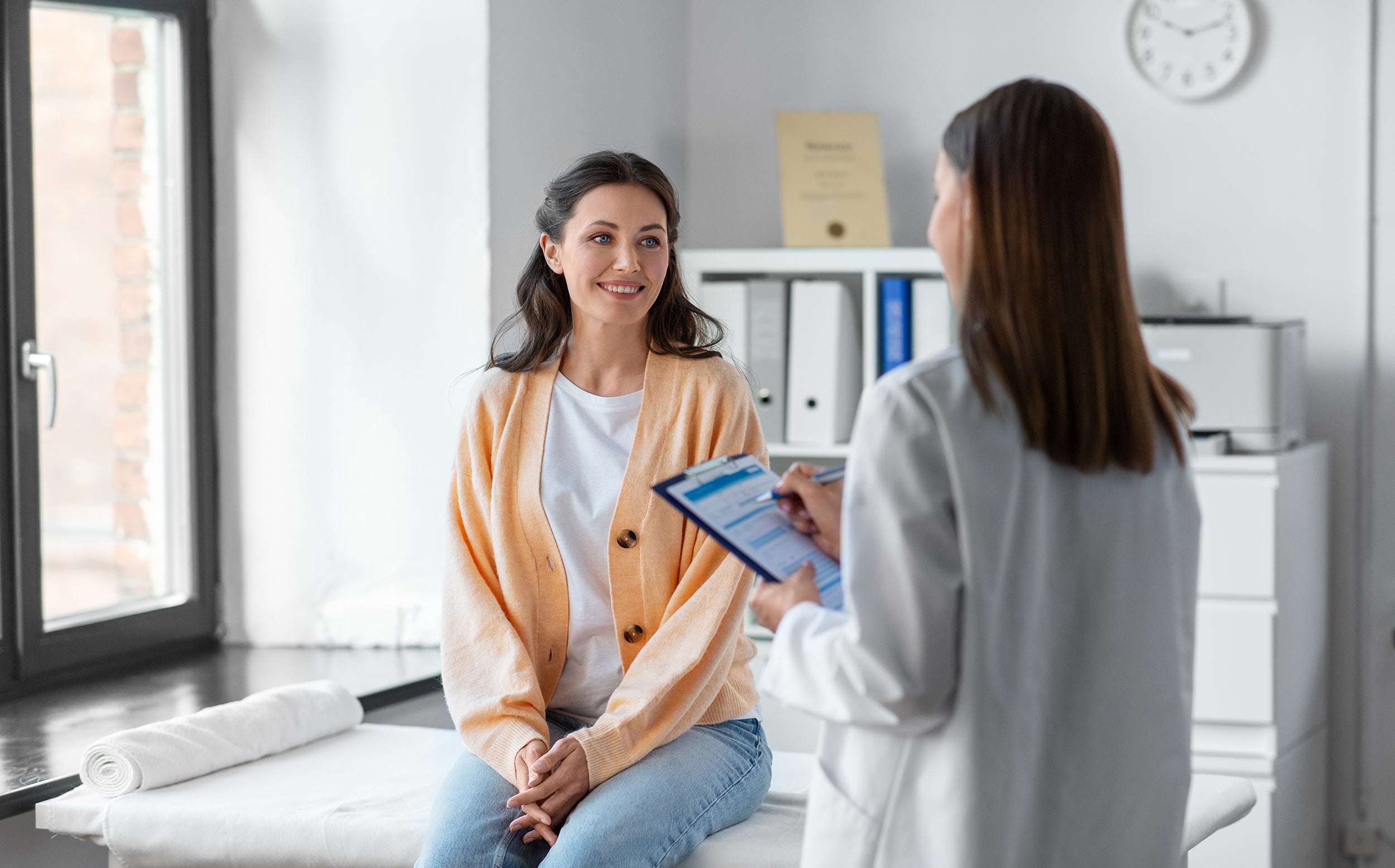 A woman sits on an exam table, smiling at a doctor who is holding a clipboard and talking to her in a bright medical office.