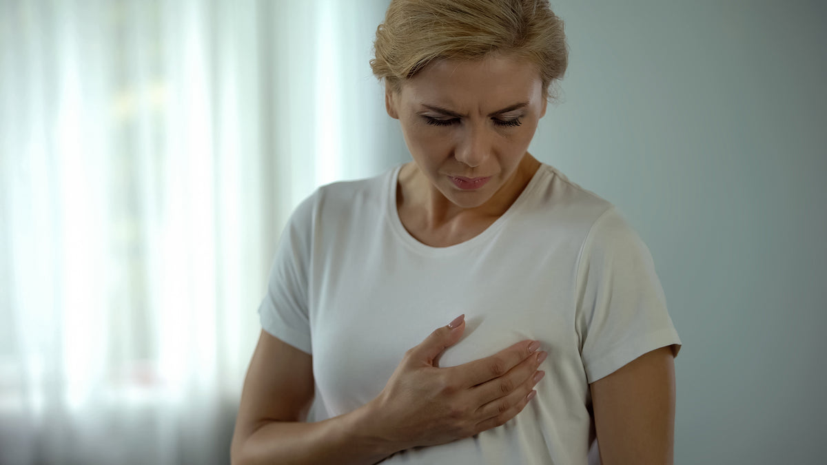 A woman in a white t-shirt holds her hand to her chest and looks down with a concerned expression, suggesting she may be experiencing chest pain or discomfort.