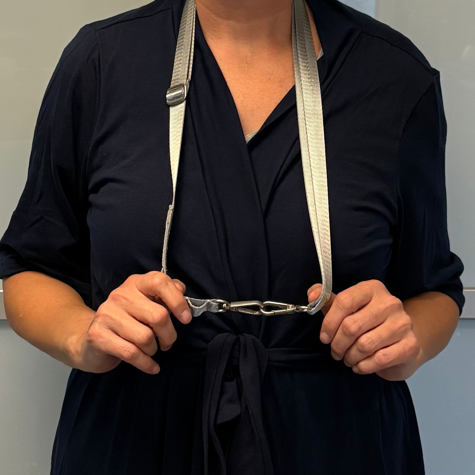 A person in a dark navy blue wrap dress holds the Sleep Again Surgical Drain Holder, a gray adjustable strap with metal hooks, looped around their neck.
