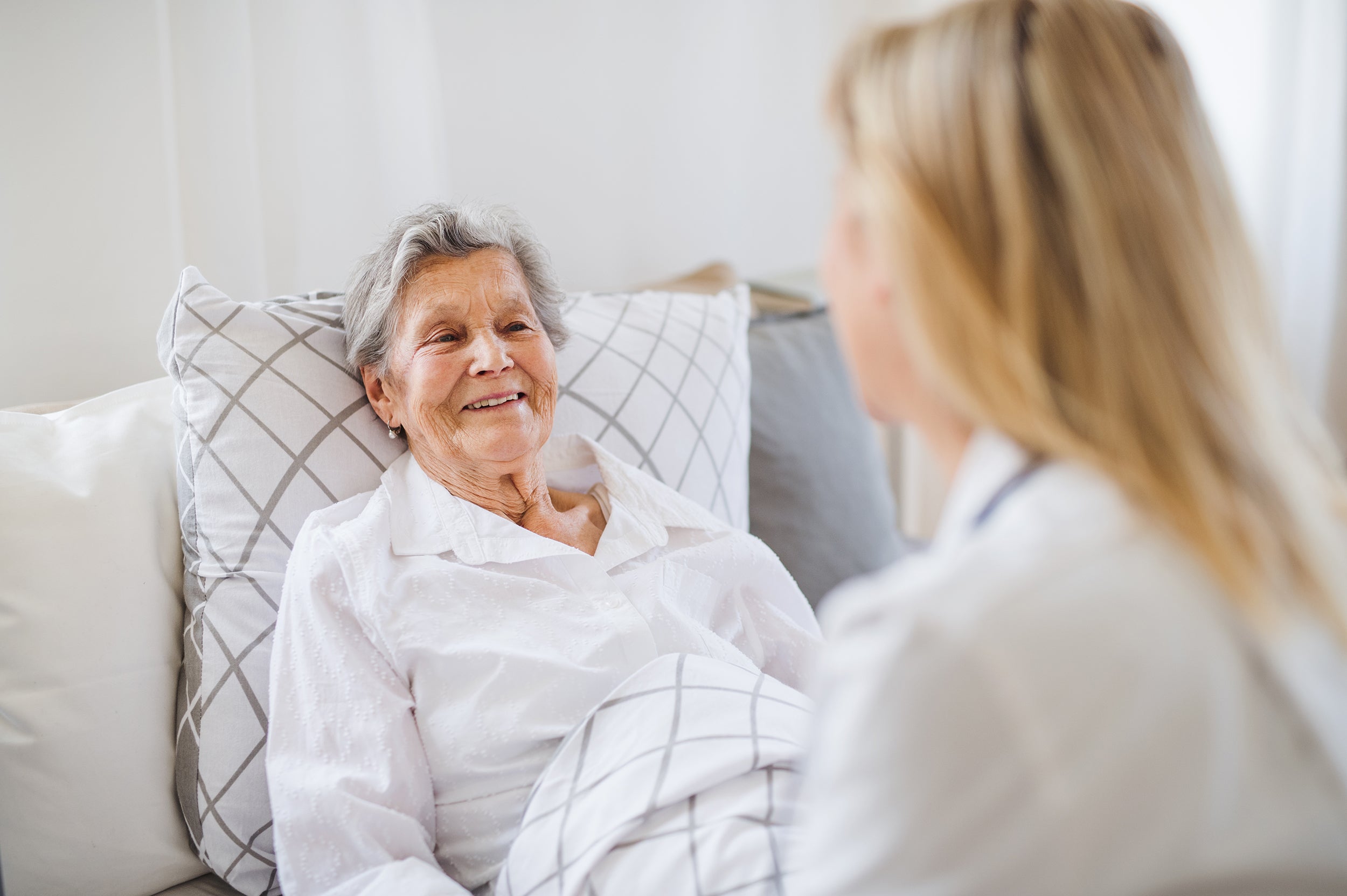 An elderly woman lies in bed, smiling while talking to a blonde woman sitting beside her, suggesting a caring or comforting interaction in a home or healthcare setting.
