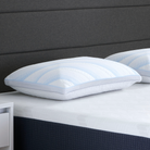 The Comfort Tech Dual Comfort Foam Pillow with light blue stripes rests on a neatly made bed with a white mattress, featuring a dark upholstered headboard and a partially visible small white nightstand on the left.