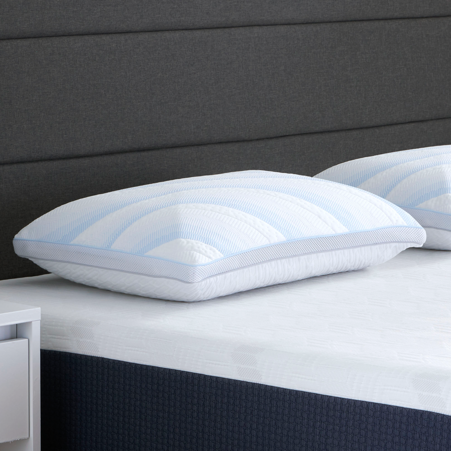 The Comfort Tech Dual Comfort Foam Pillow with light blue stripes rests on a neatly made bed with a white mattress, featuring a dark upholstered headboard and a partially visible small white nightstand on the left.