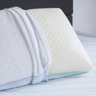A close-up of the Comfort Tech Dual Comfort Foam Pillow with its removable cover partially unzipped, revealing the perforated foam inside, resting on a light blue striped bed sheet.