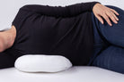 A person lies on their side with a Side Sleeping Chest Pillow under their waist, wearing a black long-sleeve shirt and blue jeans, set against a plain white background.