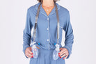 A woman wearing blue pajamas and a heart necklace stands against a white background, holding two surgical drain bulbs secured in the Sleep Again Surgical Drain Holder around her neck for post-operative drain management.