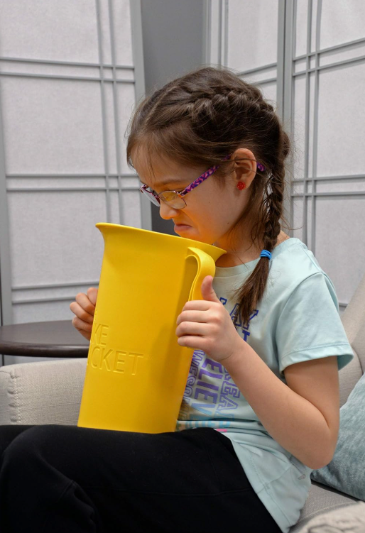 A young girl with braided hair and glasses grimaces while holding The Puke Bucket (3.0L) – a reusable container for vomiting – close to her mouth as she sits on a couch.