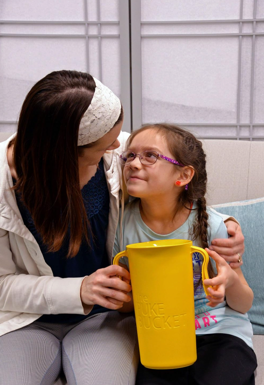 A woman comforts a young girl holding The Puke Bucket (3.0L) – a reusable container for vomiting – which is large and yellow. The girl, who wears glasses, smiles up at the woman as she puts an arm around her. They are seated together on a light-colored couch indoors.