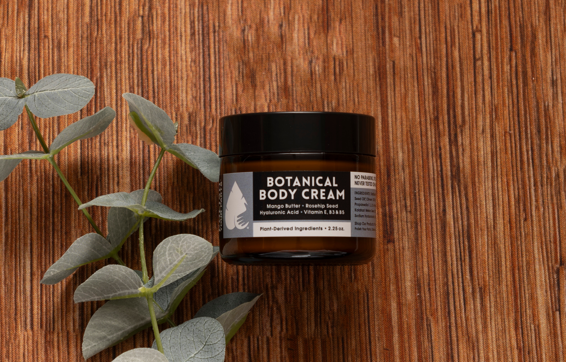 A dark jar labeled "Premium Soothing Skin & Lips" rests on a wooden surface with eucalyptus leaves—perfect for a cancer care gift box. The label highlights mango butter, rosehip seed, hyaluronic acid, vitamin E, and plant-based ingredients.