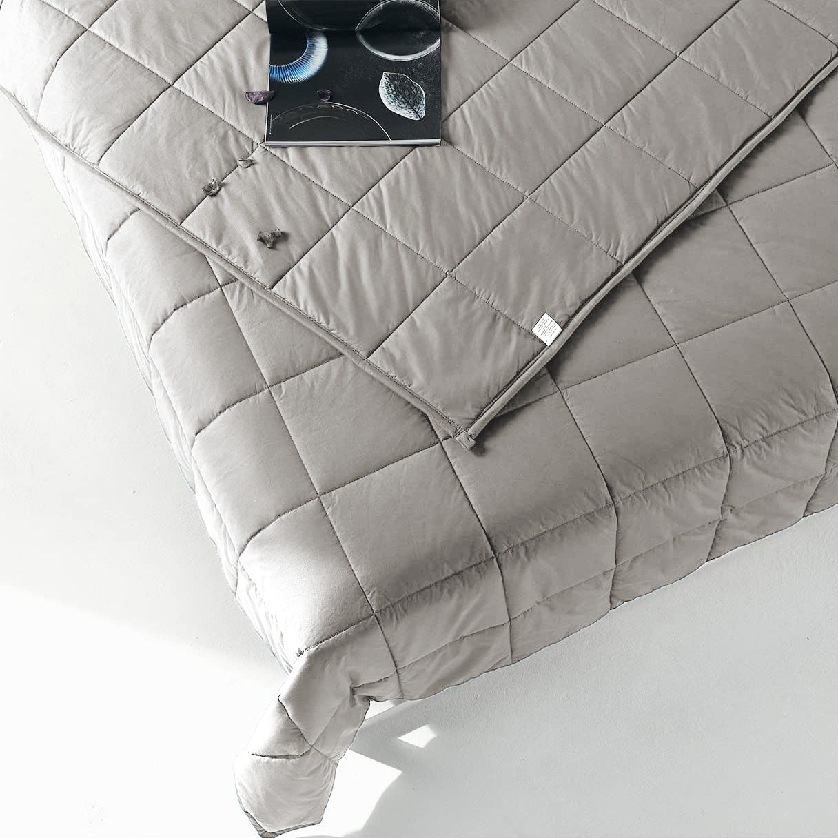 A minimal, modern bed features the Pillowtex Weighted Blanket in light grey, quilted and filled with glass beads. An open book and eyeglasses rest on the corner of the neatly made bed.