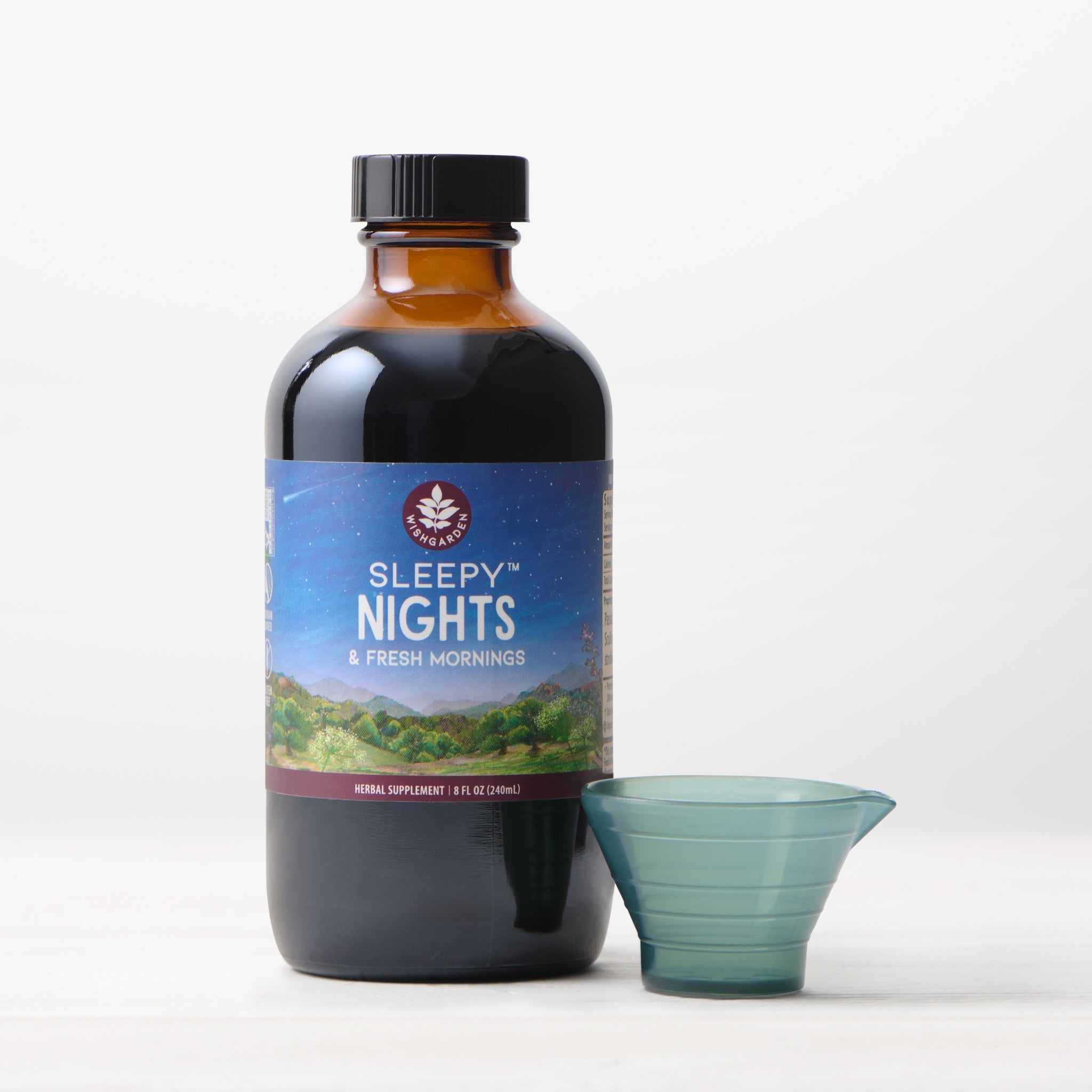A brown bottle of Sleepy Nights & Fresh Mornings organic sleep supplement, featuring a night sky and green landscape on the label, stands beside a small blue plastic dosage cup on a white surface.