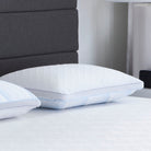 Two Comfort Tech Dual Comfort Foam Pillows with textured white covers rest on a neatly made bed with a white sheet, while a dark gray upholstered headboard and part of a nightstand with a lamp are visible in the background.