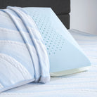 A Comfort Tech Dual Comfort Foam Pillow with a blue cover sits upright on a white and light blue striped bed, featuring small ventilation holes and a textured pattern, with a matching blanket draped over the cooling pillow.