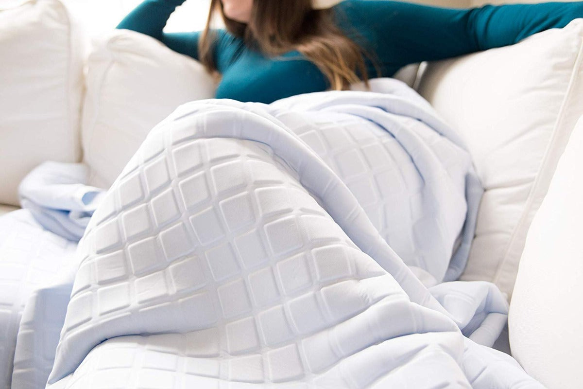 A woman in a teal long-sleeve shirt relaxes on a white couch, covered with the Final Sale: Opulence Glacier Blanket, a cooling and weighted white blanket with a geometric square pattern. Her face is partially out of frame.