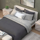 A neatly made bed with gray and light beige bedding, featuring the PureCare Duvet Cover | Soft Touch Bamboo, multiple pillows, a beige upholstered headboard, and two wooden nightstands in a modern bedroom with a green wall and abstract art.